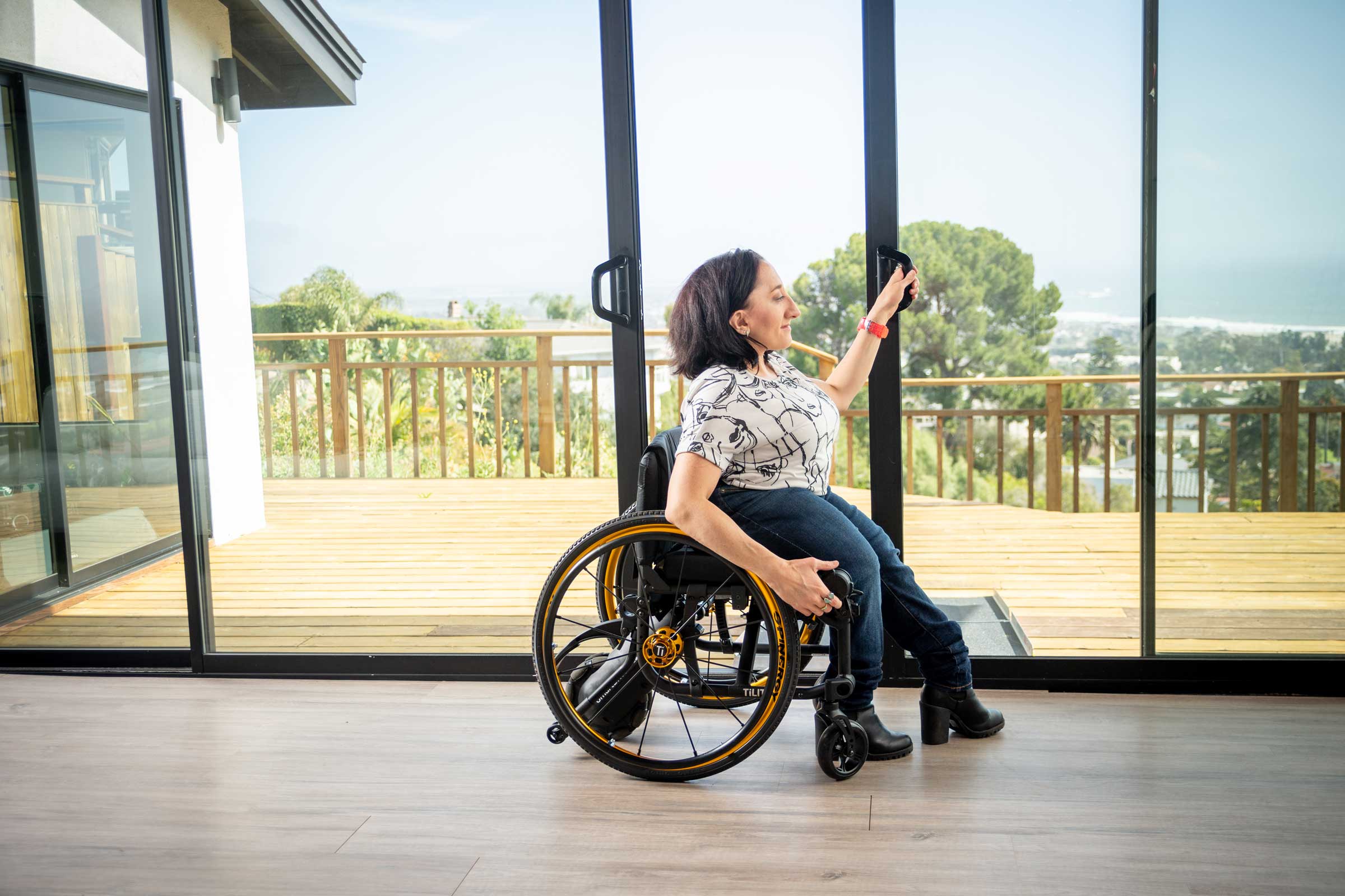 Side image of a woman in a beautiful house riding the SmartDrive MX2+ Power Assist for Manual Wheelchairs. Discreet and easy-to-use electric motor that attaches to the back of a manual wheelchair.