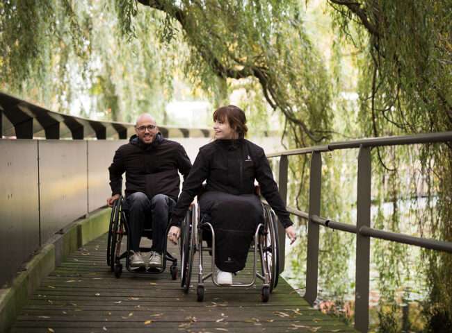 Image of a man and woman riding happily in their black Raindek Parka's - Waterproof, insulated wheelchair cover for lower body, providing warmth and protection from the elements.